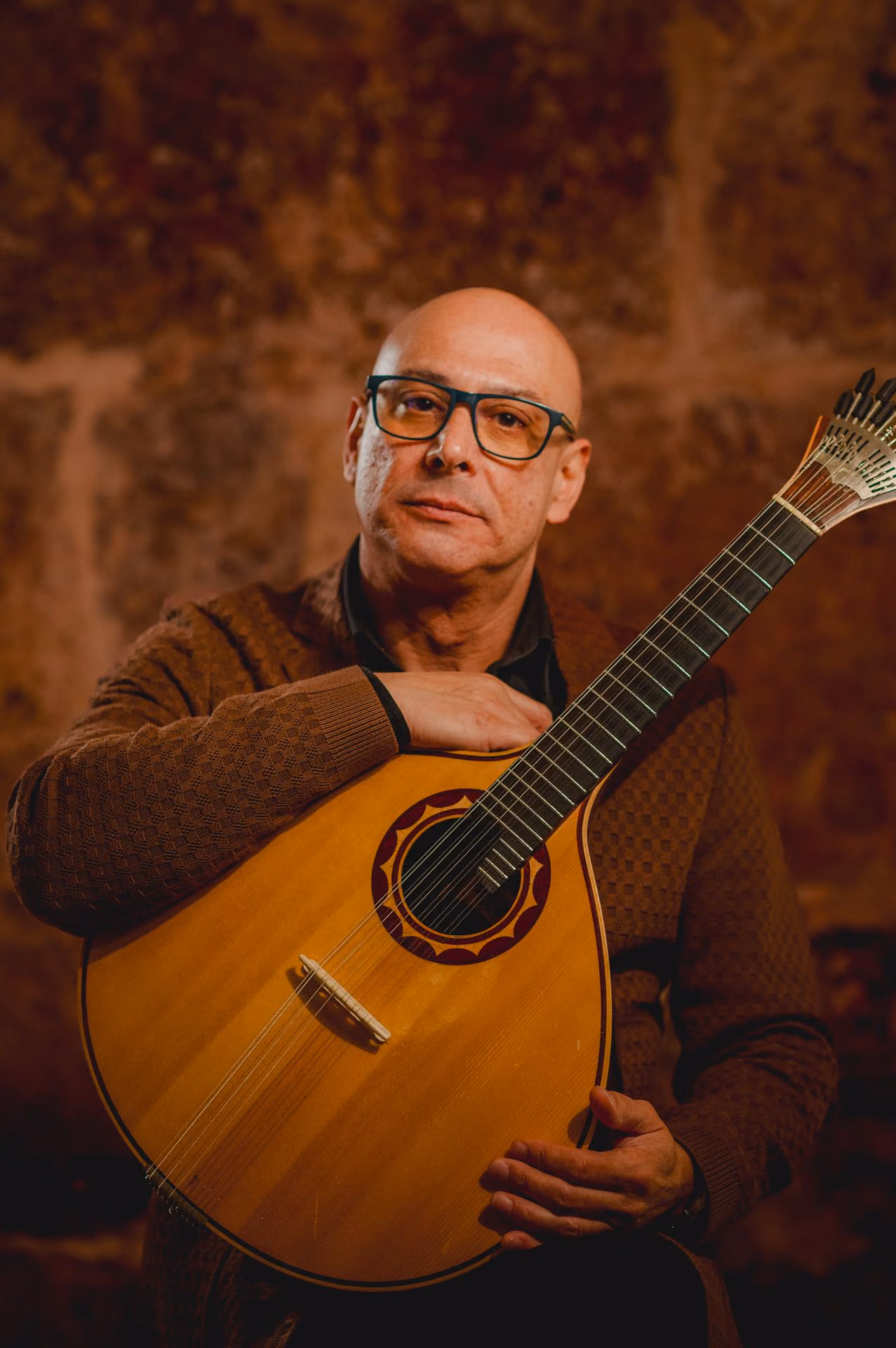 De pie, un guitarrista calvo con gafas y chaqueta marrón sostiene una guitarra portuguesa de doce cuerdas, la guitarra del Fado, y mira a la cámara en la sala de Fado de la Sé, en Lisboa
