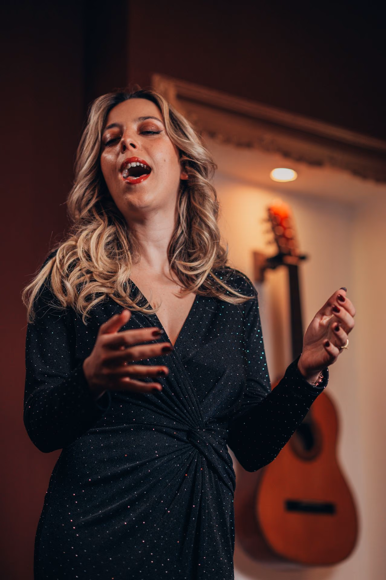 Una cantante rubia de vestido negro canta un Fado con las manos abiertas; detrás, una guitarra cuelga en una hornacina iluminada de la sala de la Baixa, en Lisboa