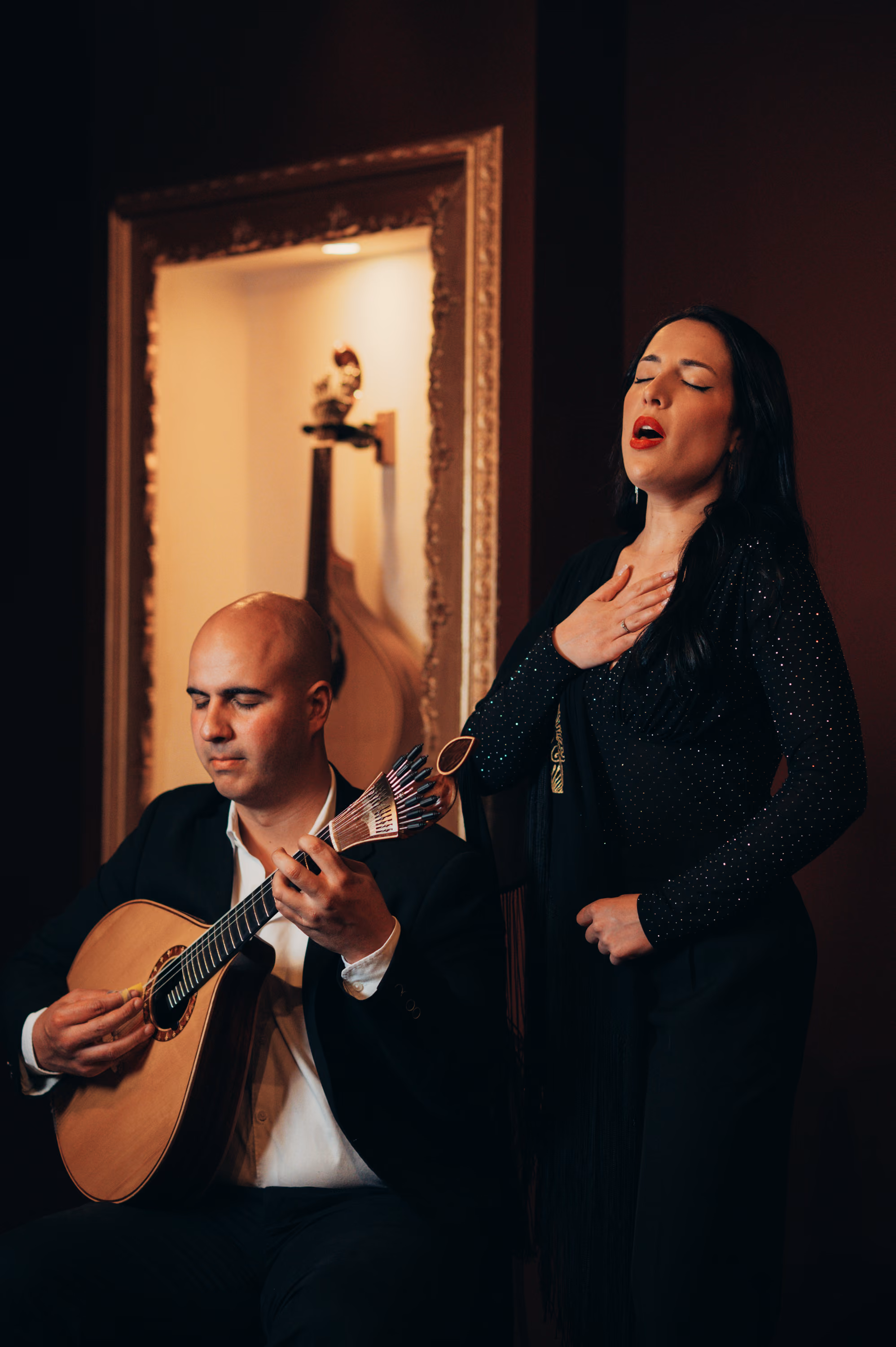 Cantante de Fado de pelo negro, con la mano en el pecho y los ojos cerrados, junto a un guitarrista sentado que toca la guitarra portuguesa; al fondo, una guitarra en una hornacina dorada de la sala de la Baixa, en Lisboa