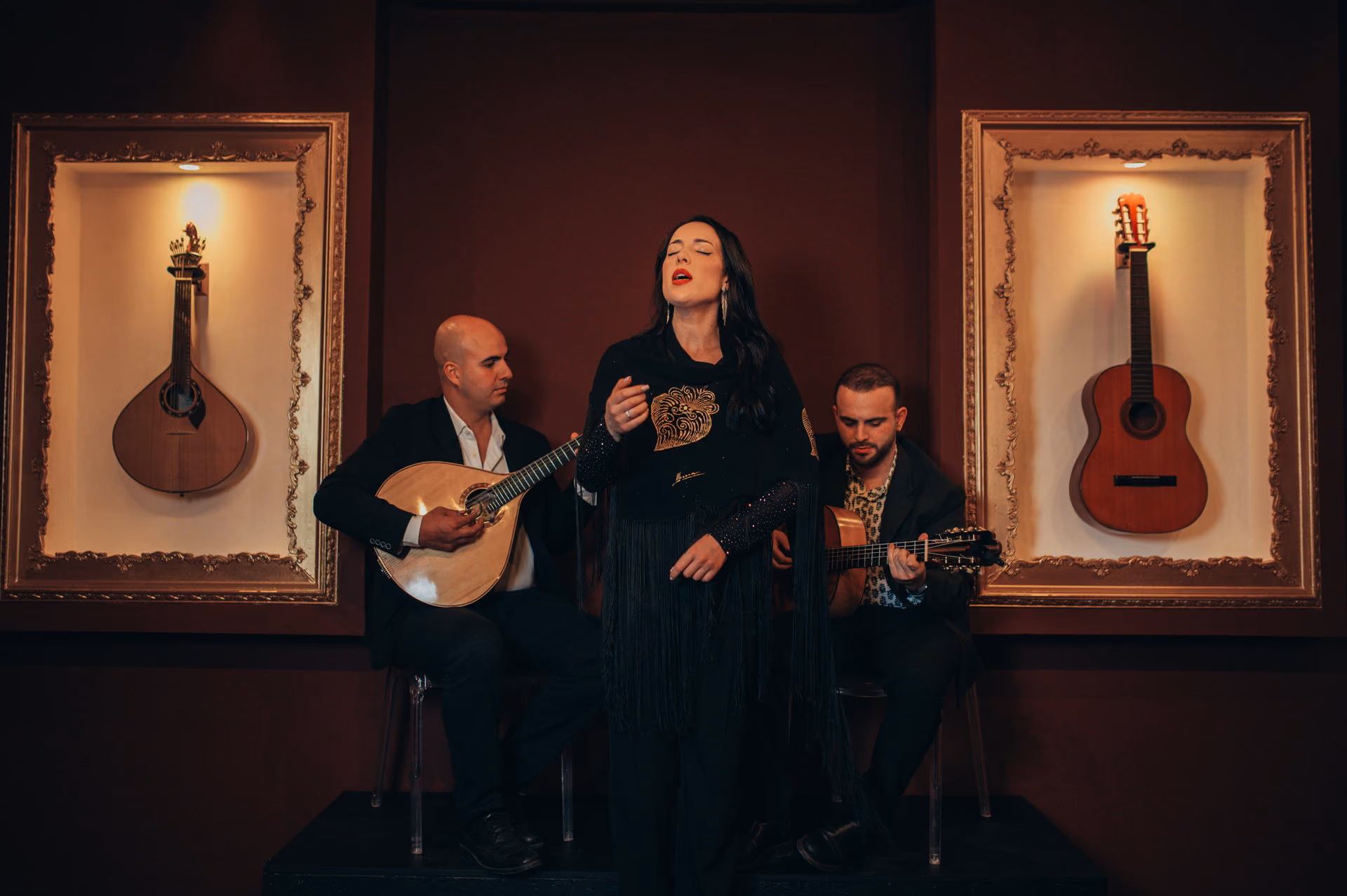 En el pequeño escenario de la Baixa, en Lisboa, una cantante de Fado de pie entre dos guitarristas sentados, uno con guitarra portuguesa y otro con guitarra clásica, con una guitarra en marco dorado a cada lado sobre la pared granate
