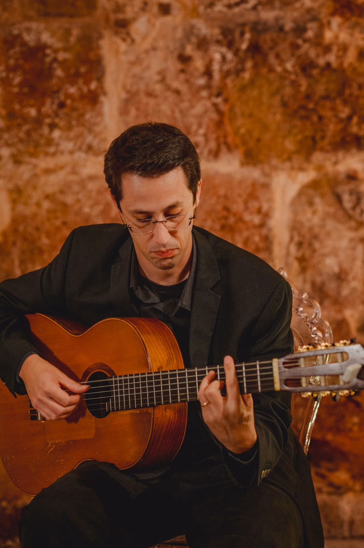 Un guitarrista con gafas, la vista en las cuerdas, toca la guitarra clásica en un concierto de Fado ante la pared de piedra de nuestra sala de la Sé, en Lisboa