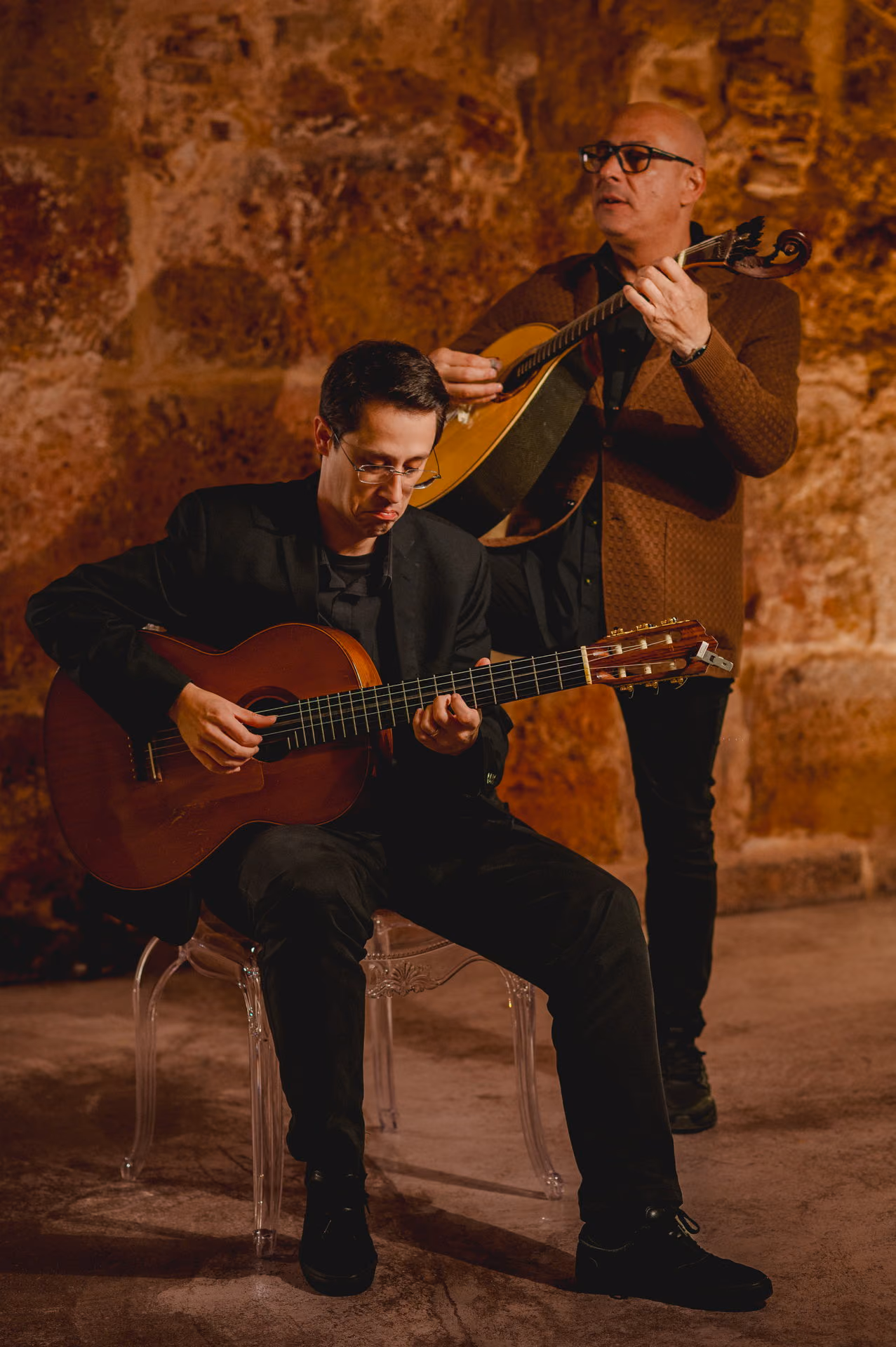 Delante, la guitarra clásica en manos de un músico sentado; detrás, de pie, la guitarra portuguesa; al fondo, la piedra antigua de la sala de Fado de la Sé, en Lisboa