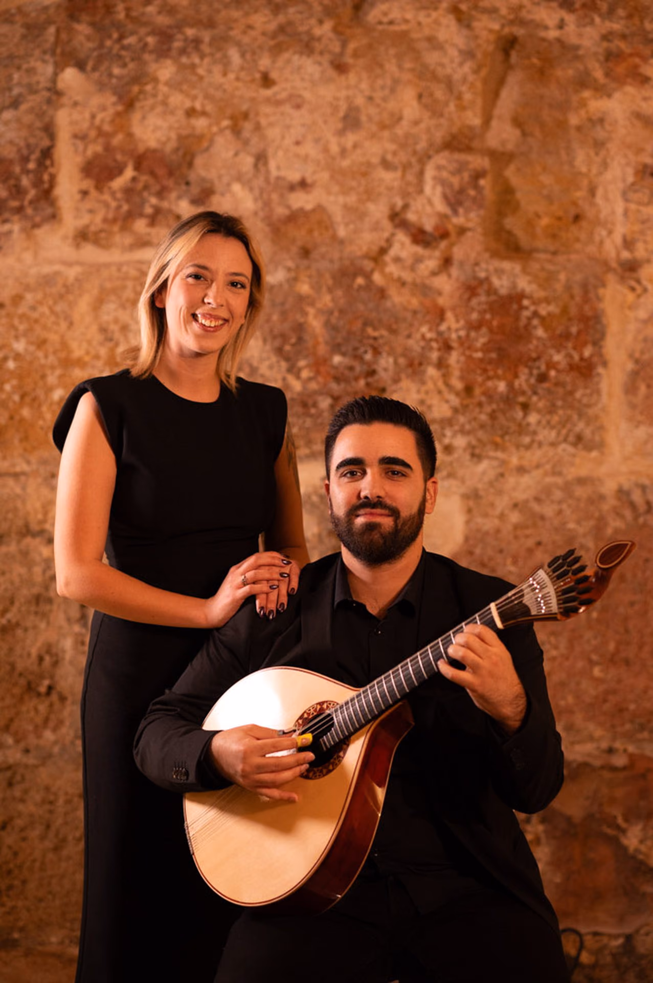 Una cantante de Fado rubia sonríe con la mano en el hombro de un guitarrista sentado con su guitarra portuguesa, ante el muro de piedra de la Sé, en Lisboa