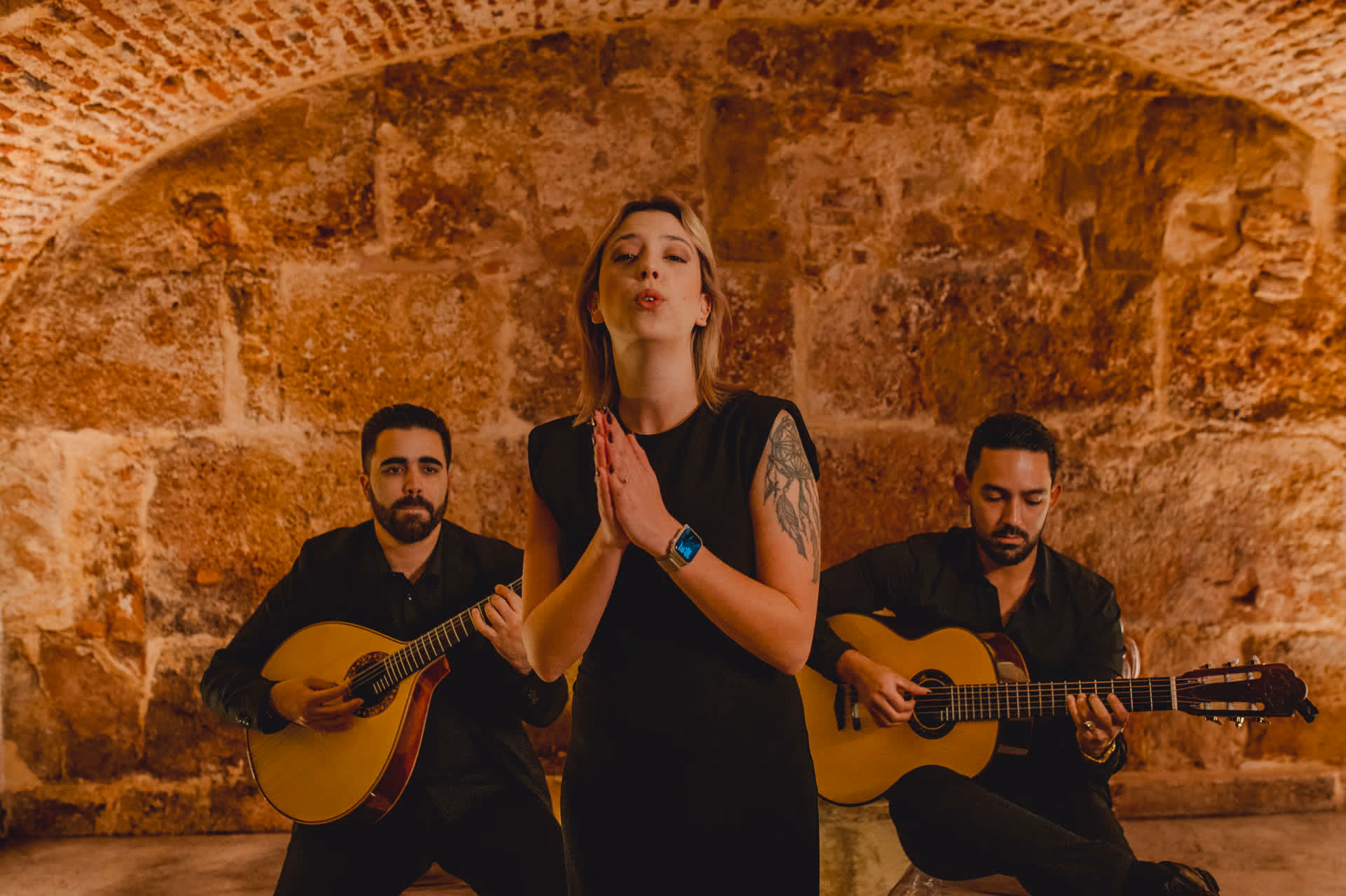 Con las manos juntas y los ojos cerrados, una cantante interpreta un Fado; detrás de ella tocan los dos guitarristas, bajo la bóveda de piedra de la Sé, en Lisboa