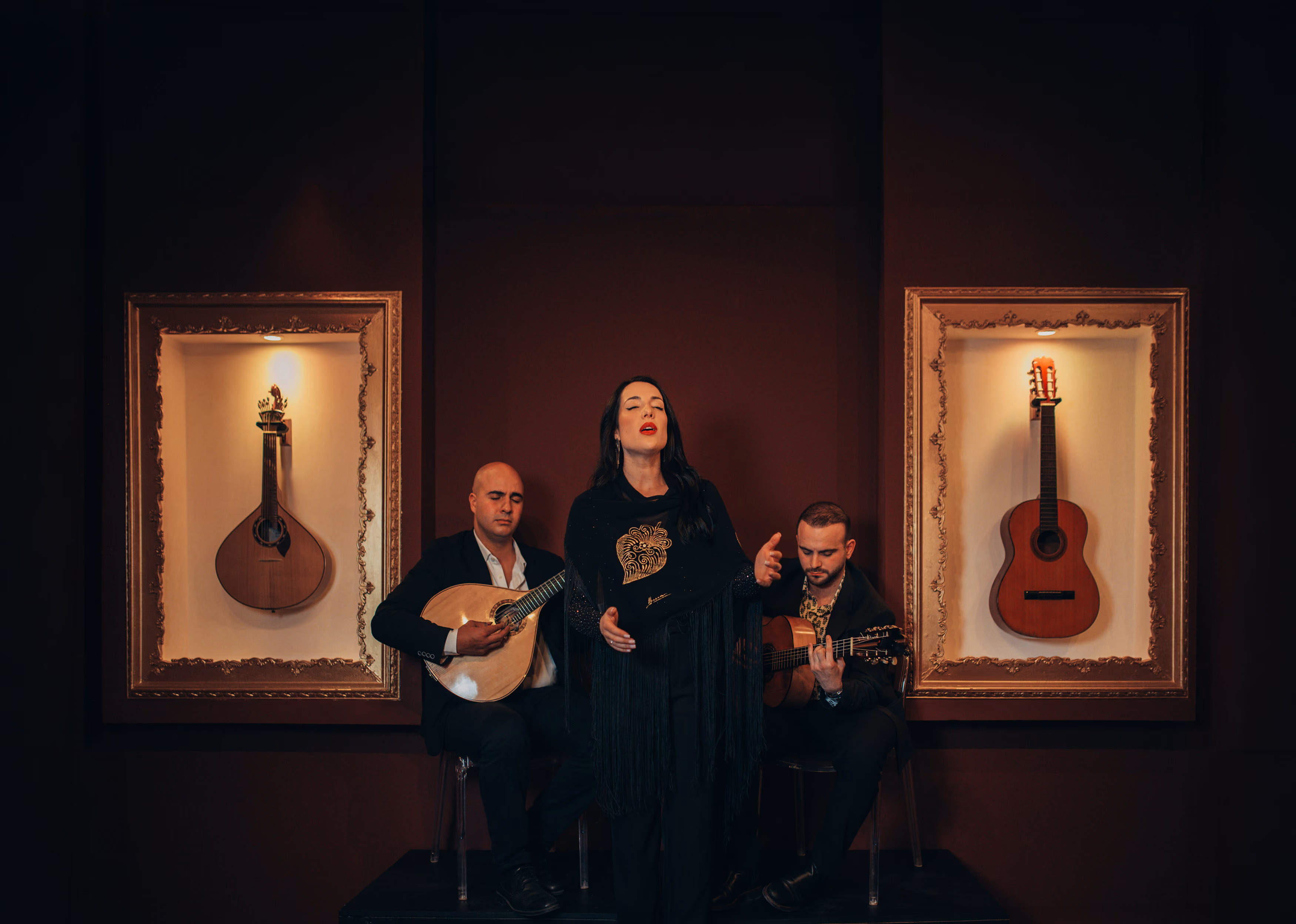 Una cantante de Fado entre dos guitarristas, uno con guitarra portuguesa y otro con guitarra clásica, en la sala de la Baixa de Lisboa; a cada lado, una guitarra en marco dorado sobre la pared color vino